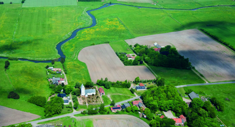 Hvid landsbykirke set fra luften, med agerlandskab og å omrkring