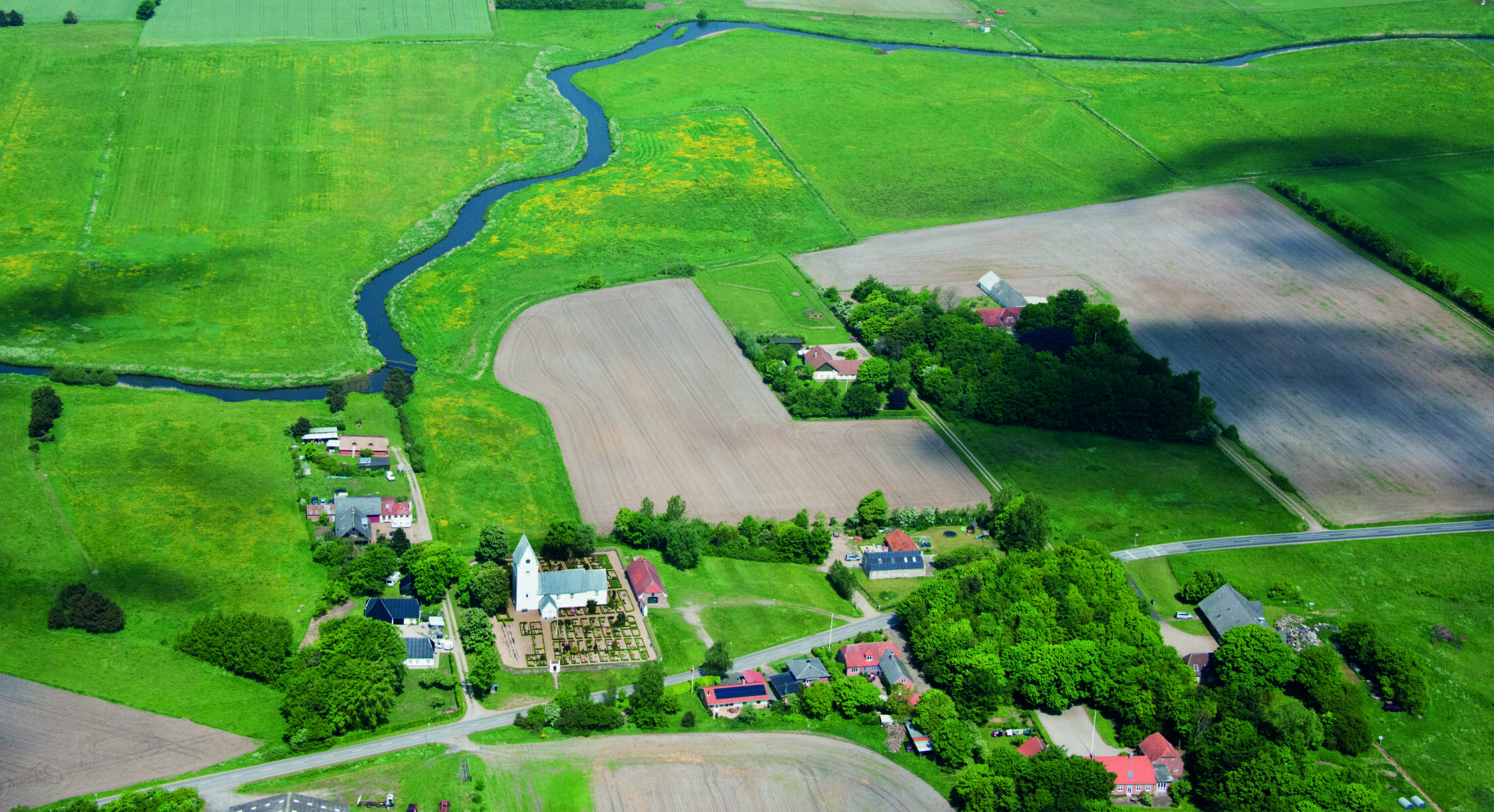 Hvid landsbykirke set fra luften, med agerlandskab og å omrkring